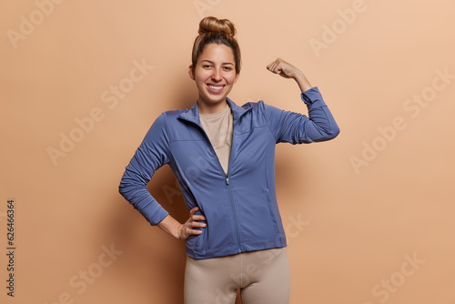 Sporty woman with hair bun feels strong shows biceps keeps arm raised up being in good physical shape dressed in sportswear demonstrates results after everyday workout isolated over brown background