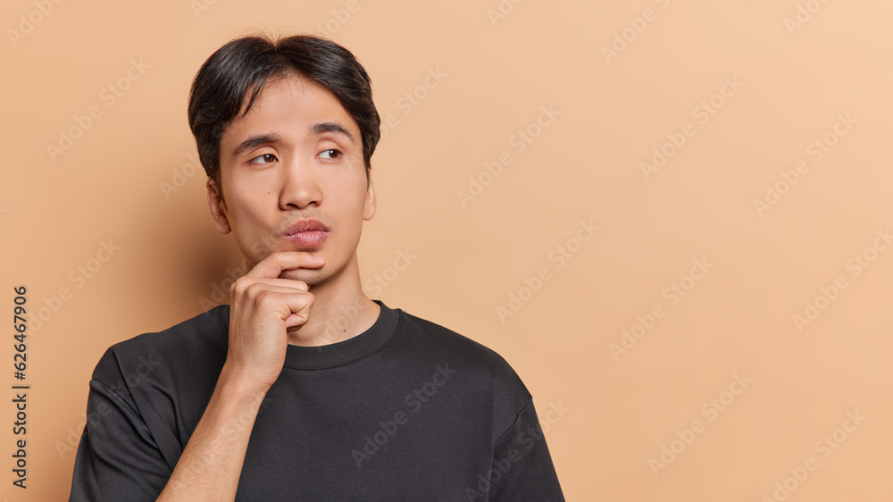 People emotions concept. Young thoughtful Asian man holding left hand on chin trying to solve difficult task or problem standing on left isolated on beige background with blank space for advertisement People emotions concept. Young thoughtful Asian man holding left hand on chin trying to solve difficult task or problem standing on left isolated on beige background with blank space for advertisement