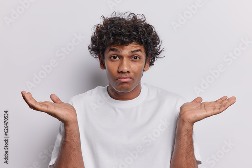 Clueless Hindu man with curly hair has raised palms and shrugged shoulders signify his lack of understanding demonstrates bewilderment and uncertainty wears basic t shirt isolated on white background