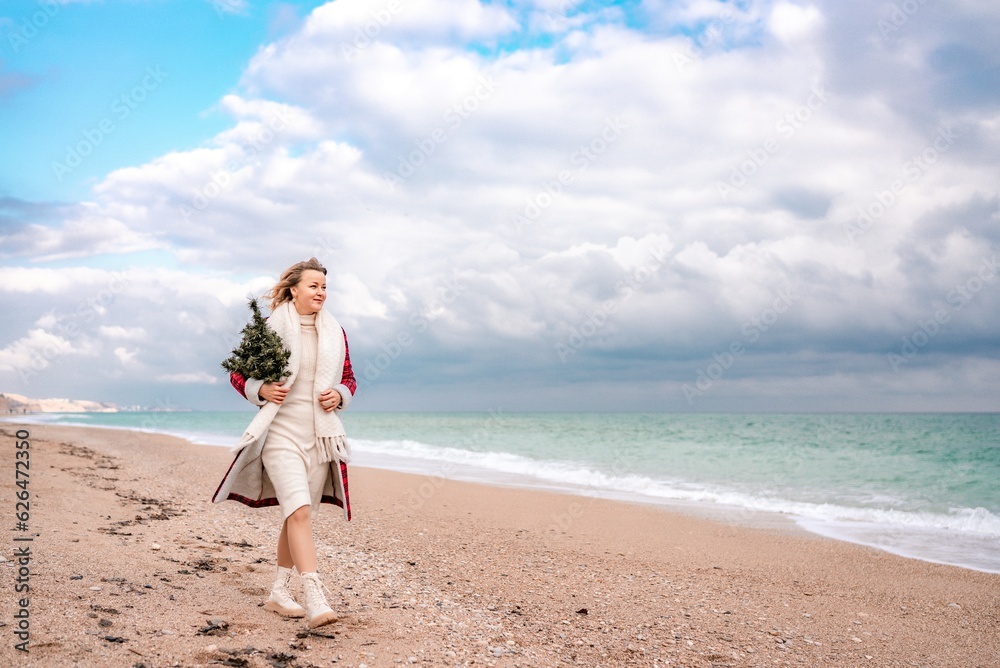 Blond woman holding Christmas tree by the sea. Christmas portrait of a happy woman walking along the beach and holding a Christmas tree in her hands. Dressed in a red coat, white dress. Blond woman holding Christmas tree by the sea. Christmas portrait of a happy woman walking along the beach and holding a Christmas tree in her hands. Dressed in a red coat, white dress.