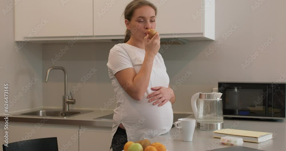 Pregnant woman standing in the kitchen and eating an apple, An apple a day keeps a doctor away Slow motion health care, wellness Pregnant woman standing in the kitchen and eating an apple, An apple a day keeps a doctor away Slow motion health care, wellness