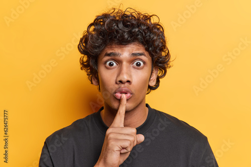 Indoor photo of young Hindu standing isolated in centre on yellow background wearing casual black tshirt keeping finger near lip making gesture to calm down be still and not say anything loudly photo