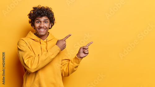 Positive emotions concept. Studio waist up of young cheerful smiling broadly Hindu guy wearing hoodie standing on left isolated on yellow background pointing at blank space for your advertisement