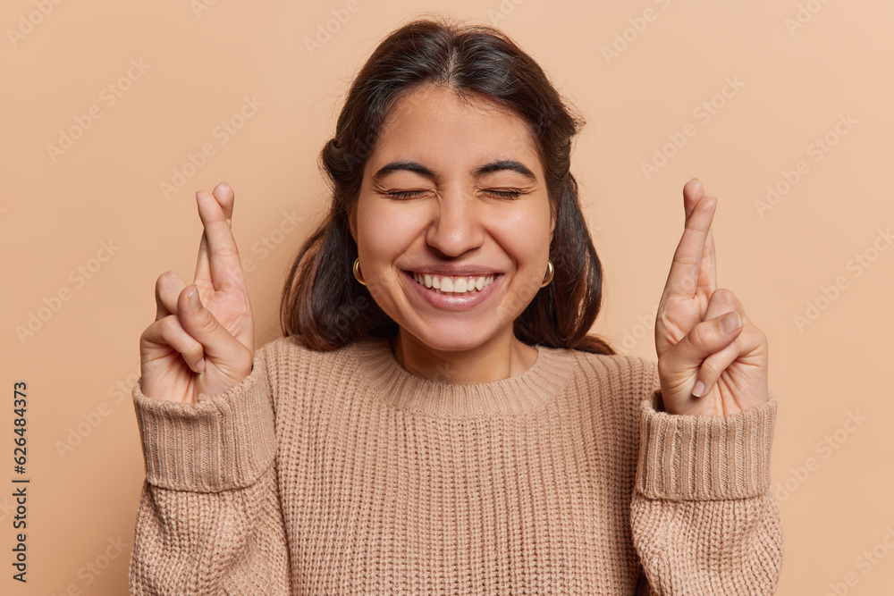Glad dark haired Iranian woman keeps her fingers adorably crossed in gesture of hope and anticipation believes in magic of making wishes come true dressed in knitted jumper isolated over brown wall
