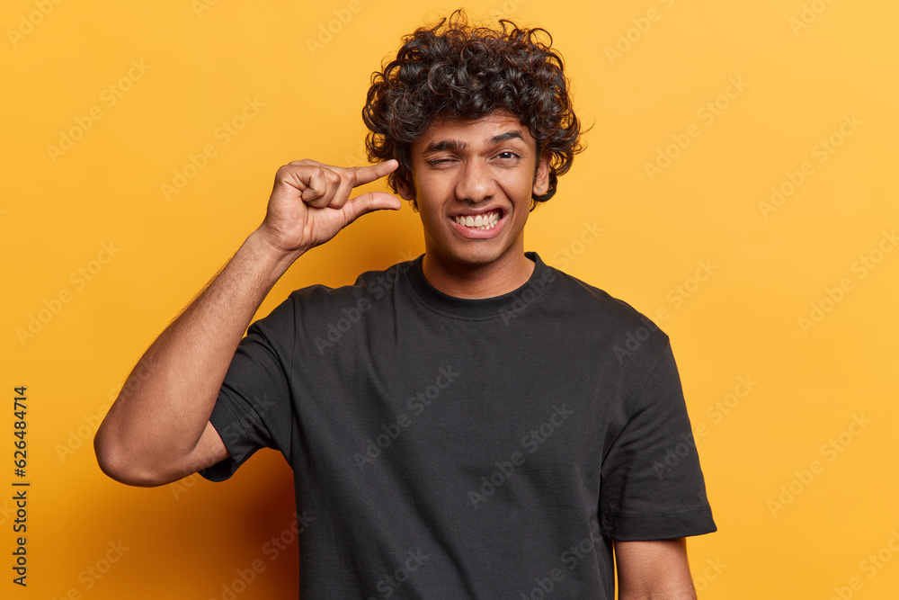 People and emotions. Studio waist up of young cheerful smiling broadly Hindu male standing in centre isolated on yellow background wearing black tshirt showing something small with left hand People and emotions. Studio waist up of young cheerful smiling broadly Hindu male standing in centre isolated on yellow background wearing black tshirt showing something small with left hand