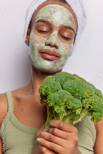 Indoor shot of calm African woman applies rejuvenating green mask to her flawless complexion uses boccoli for making natural cosmetic products wears t shirt and bath towel on head isolated on white photo