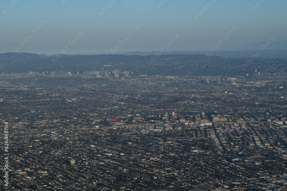 The hazy, polluted air obscures an aerial view from high above Los Angeles. The cityscape is mostly sprawling and low-rise, punctuated by a few clusters of skyscrapers, and mountains in the distance.