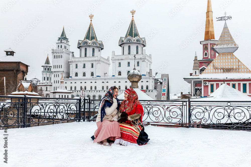 Obraz premium Two beautiful Slavic women in national traditional Slavic costumes on the roof of a wooden tower against the backdrop of a snow-covered city. Pancake day