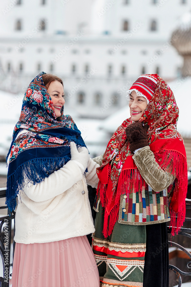 Two beautiful Slavic women in national traditional Slavic costumes on the roof of a wooden tower against the backdrop of a snow-covered city. Pancake day