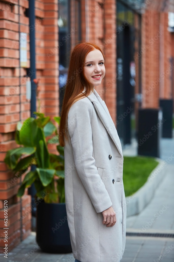 A stylish beautiful young girl is standing on a city street. Red hair. Autumn or spring season.