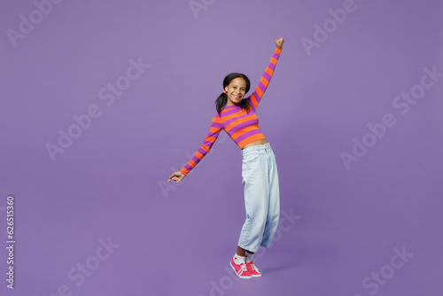 Full body little kid teen girl 15-16 years old wear striped orange sweatshirt stand on toes leaning back dance with outstretched hands isolated on plain purple background. Childhood lifestyle concept.