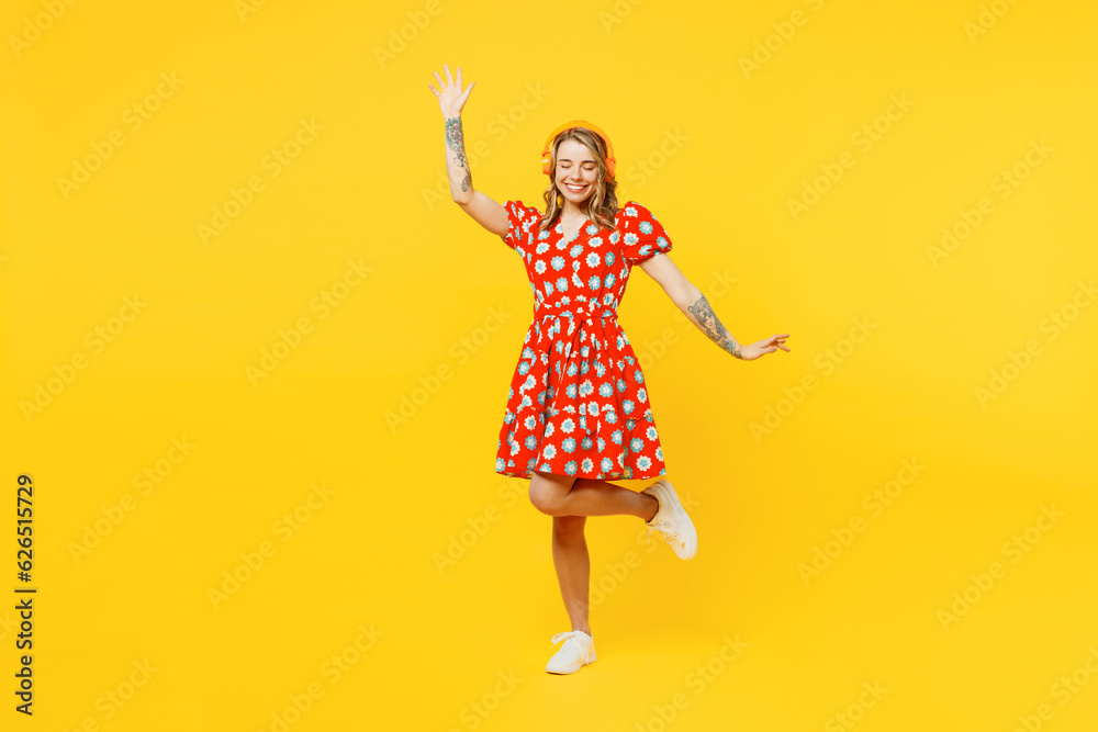Full body young smiling fun caucasian woman she wear red dress casual clothes listen to music in headphones dance raise up hands isolated on plain yellow background studio portrait. Lifestyle concept.