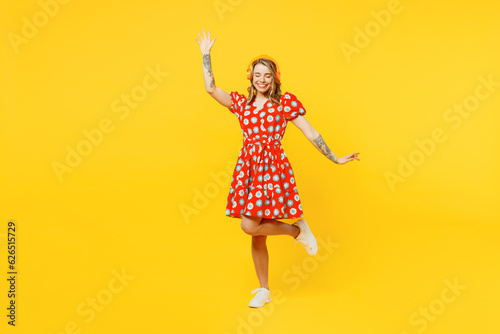 Full body young smiling fun caucasian woman she wear red dress casual clothes listen to music in headphones dance raise up hands isolated on plain yellow background studio portrait. Lifestyle concept.