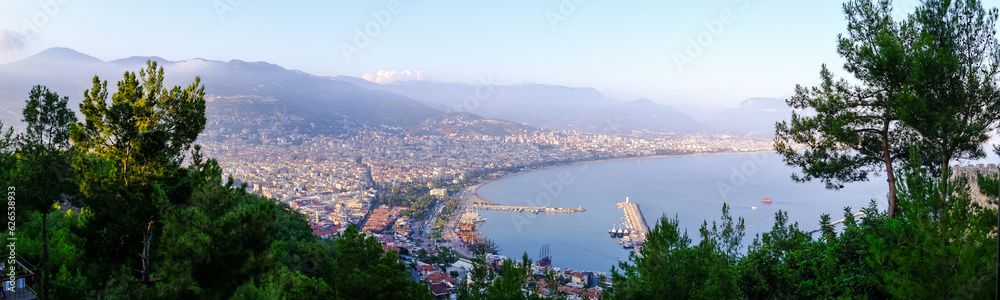 Enjoy the stunning view of Alanya from the observation deck, with lush trees, blue sea, and the iconic Lighthouse and Port of Alanya. The rocky peninsula adds a dramatic touch to the panoramic scene.