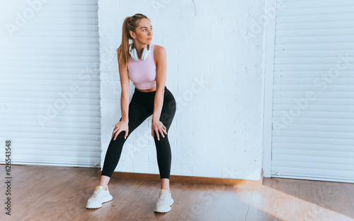 Focused caucasian girl in sportswear standing at gym leans on wall looks aside relaxing after endurance training. American young woman with headphones on neck makes break at sport club.