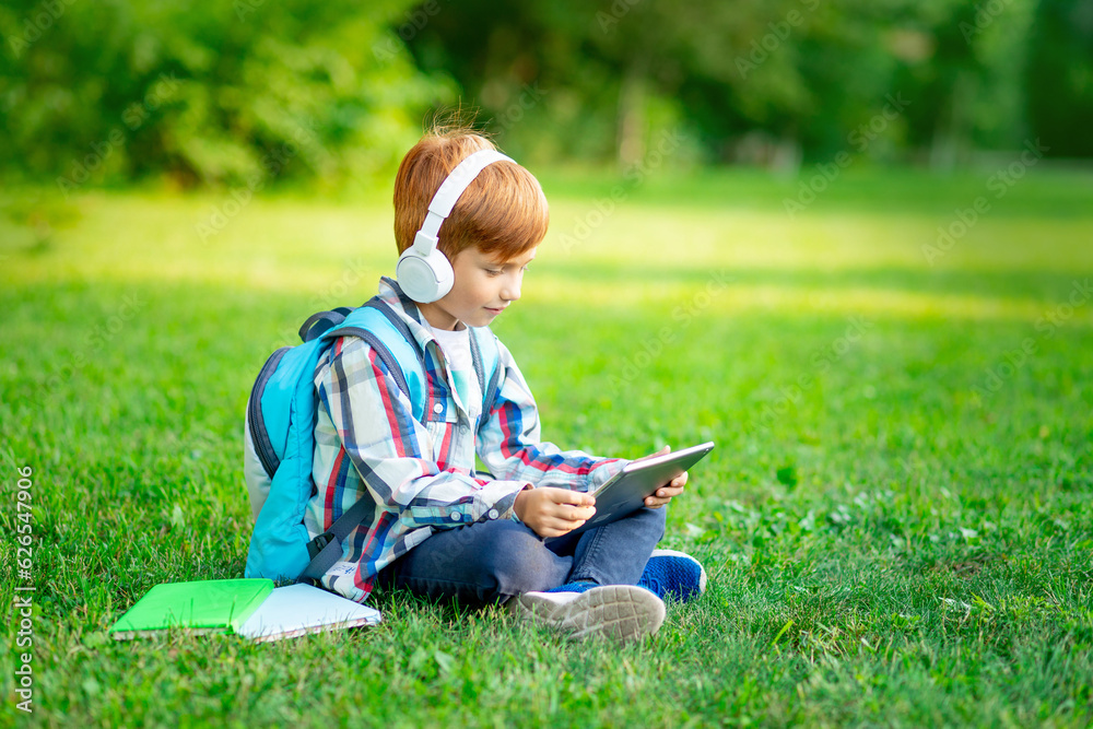 a first-grader boy with a backpack and a tablet with headphones on a green lawn reading a book or doing homework, going back to school