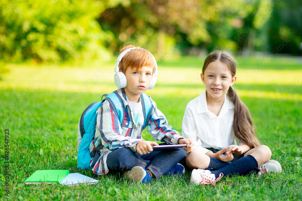 Fototapeta premium a boy and a girl with backpacks are sitting on the lawn and reading a book and a tablet with headphones, the concept is back to school, brother and sister are doing homework on the street