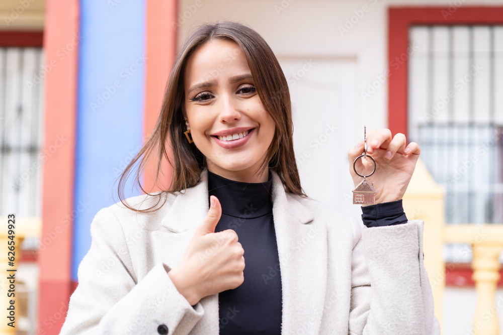 Young pretty woman holding home keys at outdoors with thumbs up because something good has happened Young pretty woman holding home keys at outdoors with thumbs up because something good has happened