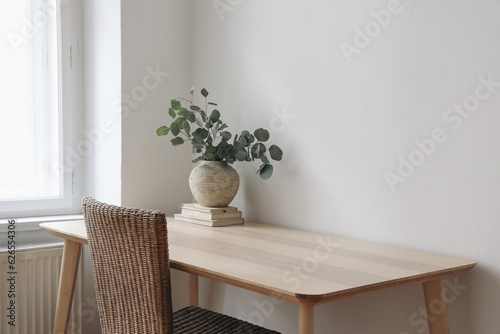 Stylish interior. Living room with vase, eucalyptus tree branches, old books on wooden table. Rattan chair near window. Minimalist concept of home decor. Scandinavian design. Blank wall template