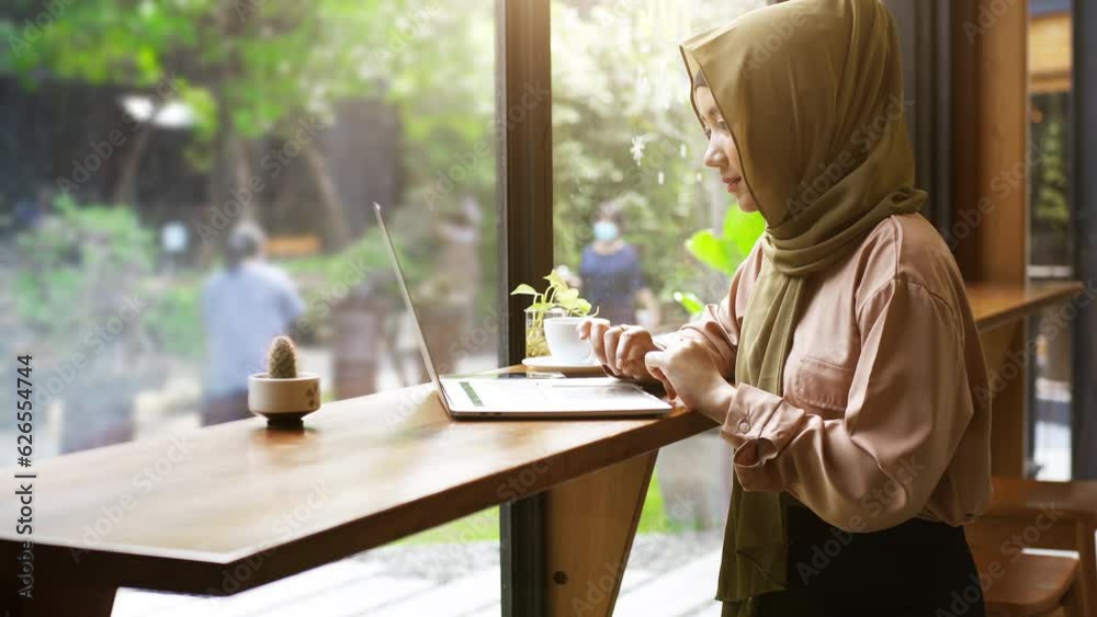 Young beautiful modern Asian Muslim women sitting by the window enjoying a relaxing moment working on a laptop in the coffee shop on a bright sunny day Young beautiful modern Asian Muslim women sitting by the window enjoying a relaxing moment working on a laptop in the coffee shop on a bright sunny day