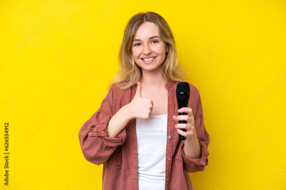 Young singer caucasian woman picking up a microphone isolated on yellow background giving a thumbs up gesture