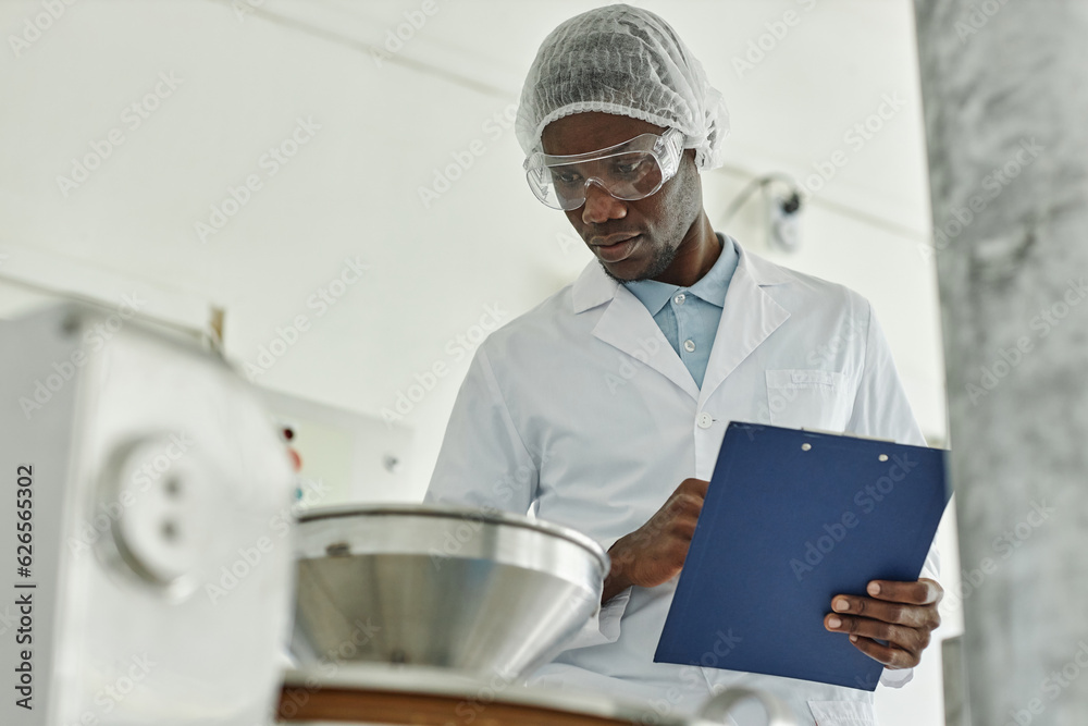 Portrait of black young man wearing lab coat and holding clipboard while overseeing production at food factory, copy space