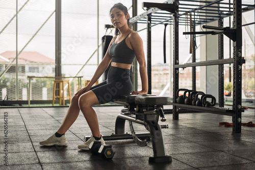 Strong Asian woman sitting on bench with dumbbell at cross fit gym. Athlete female wearing sportswear workout on grey gym background with weight and dumbbell equipment. Healthy lifestyle.