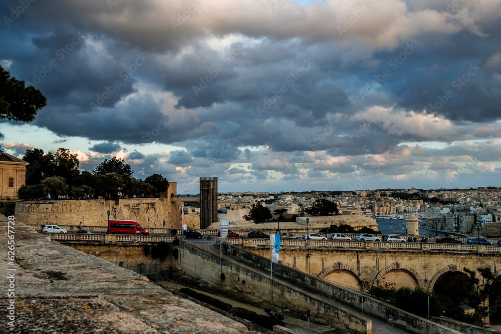 The setting sun shines through the clouds and colors everything orange. In the foreground the old town of Valletta