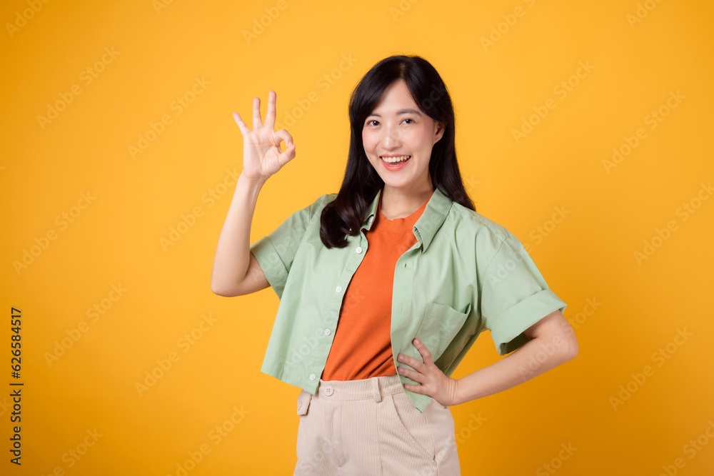 young Asian woman in her 30s, dressed in orange shirt and green jumper. Her okay hand gesture and gentle smile, isolated on a yellow background, convey a positive message through body language. young Asian woman in her 30s, dressed in orange shirt and green jumper. Her okay hand gesture and gentle smile, isolated on a yellow background, convey a positive message through body language.