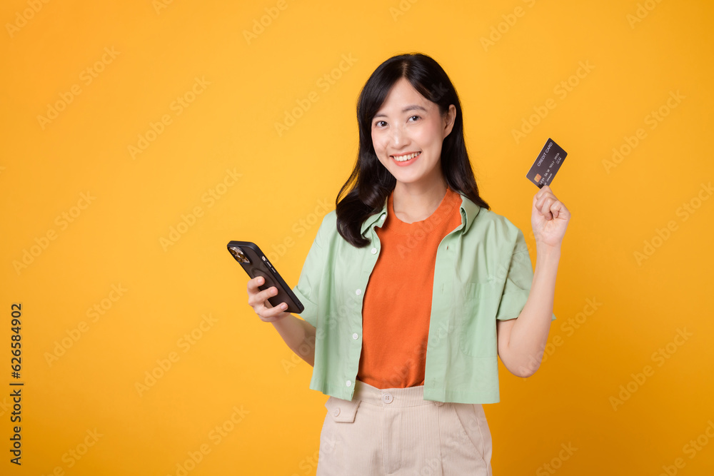 excitement of online shopping with a cheerful young Asian woman 30s, wearing orange shirt and green jumper, using smartphone while displaying credit card on yellow studio background. Mobile concept. excitement of online shopping with a cheerful young Asian woman 30s, wearing orange shirt and green jumper, using smartphone while displaying credit card on yellow studio background. Mobile concept.