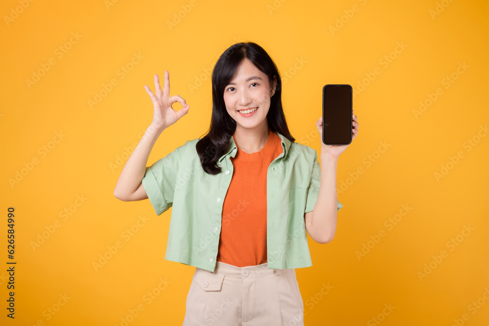 excitement of a new mobile application with a cheerful young Asian woman 30s, donning orange shirt and green jumper, displaying smartphone screen with okay hand gesture on yellow studio background. excitement of a new mobile application with a cheerful young Asian woman 30s, donning orange shirt and green jumper, displaying smartphone screen with okay hand gesture on yellow studio background.