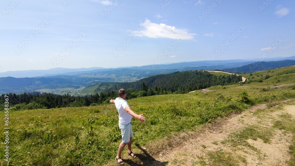 Fototapeta premium Young man standing with raised hands on top of a mountain and enjoying the view of the valley