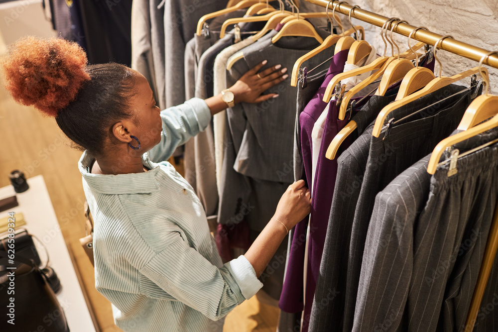 High angle portrait of African American young woman browsing clothes on rack in clothing store, copy space
