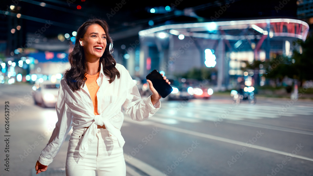 Fototapeta premium Trendy happy woman listening favorite music in earphones and dancing, walking in the evening on city street, free space