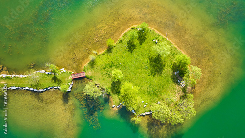 Aerial over teal green pond with small island and land bridge leading to red walking bridge