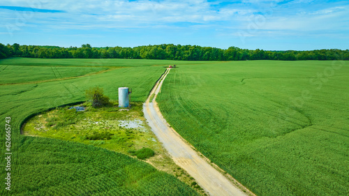 Green fields surrounding dirt road with abandoned structure and distant forest aerial