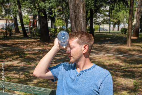 Man suffering from heat, guy with heatstroke. Caucasian male presses bottle of water to face to cool off suffering from heat wave, stuffiness