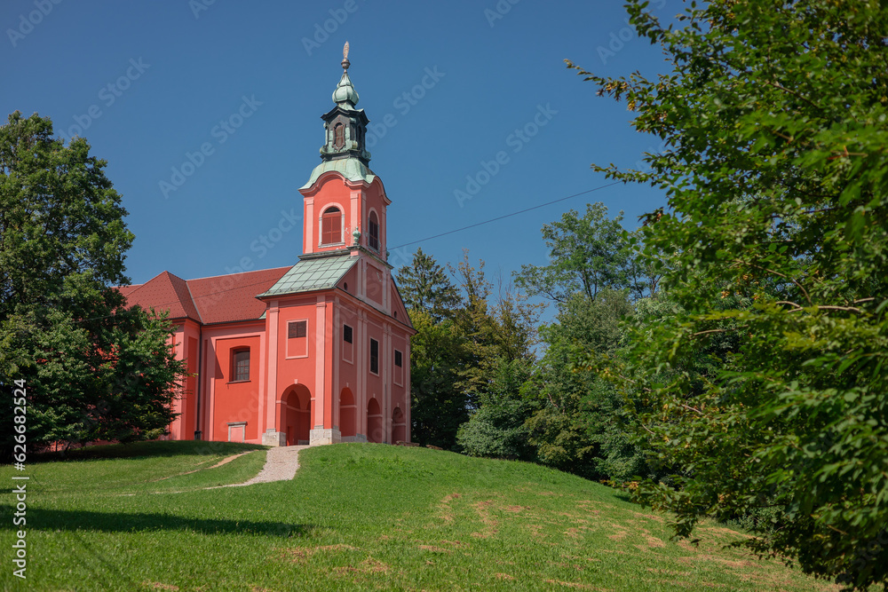 Red church on Rozik hill above Ljubljana surrounded by lush green trees and grassy meadows. Beautiful scenery around a tyoical church.