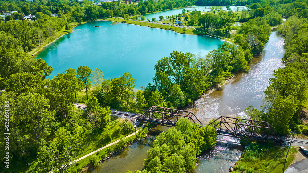 Fototapeta premium Aerial beautiful teal blue pond with summertime trees and a river running under a train bridge