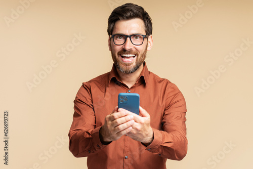 Portrait overjoyed latin man of handsome holding mobile phone shopping online isolated on background. Successful smiling hipster sports betting, win money. Emotional trader looking at camera © Maria Vitkovska
