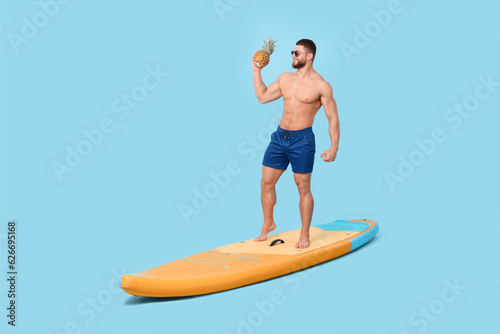Happy man with pineapple posing on SUP board against light blue background © New Africa