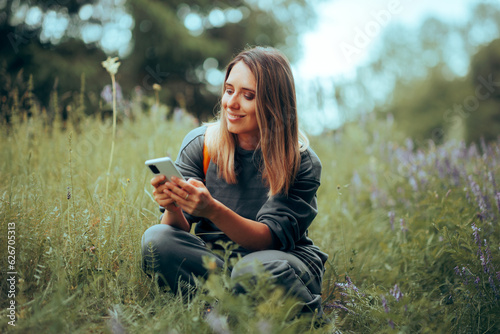 Happy Woman Checking her Phone Sitting in the Grass in Nature. Carefree girl browsing the internet enjoying natural landscape
