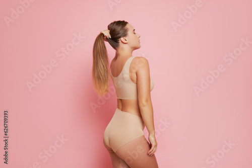 Slim half-naked girl on pink background side shot, wearing beige sports underwear,holds her long hair up, natural beauty concept, copy space.