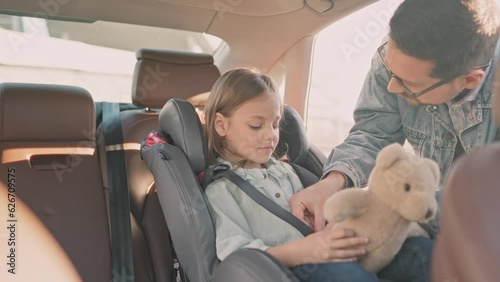Adult man securing little daughter in car seat in his vehicle. Cheerful young handsome father on trip in car with teen daughter touching her nose to cheer up. Childhood and parenting, travel concept.