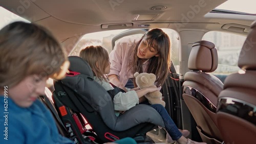 Pretty young lady ensures that two little ones are securely fastened in their seat belts. Happy family is getting set for car excursion, mother motivates children. Parent prioritize safety trip.