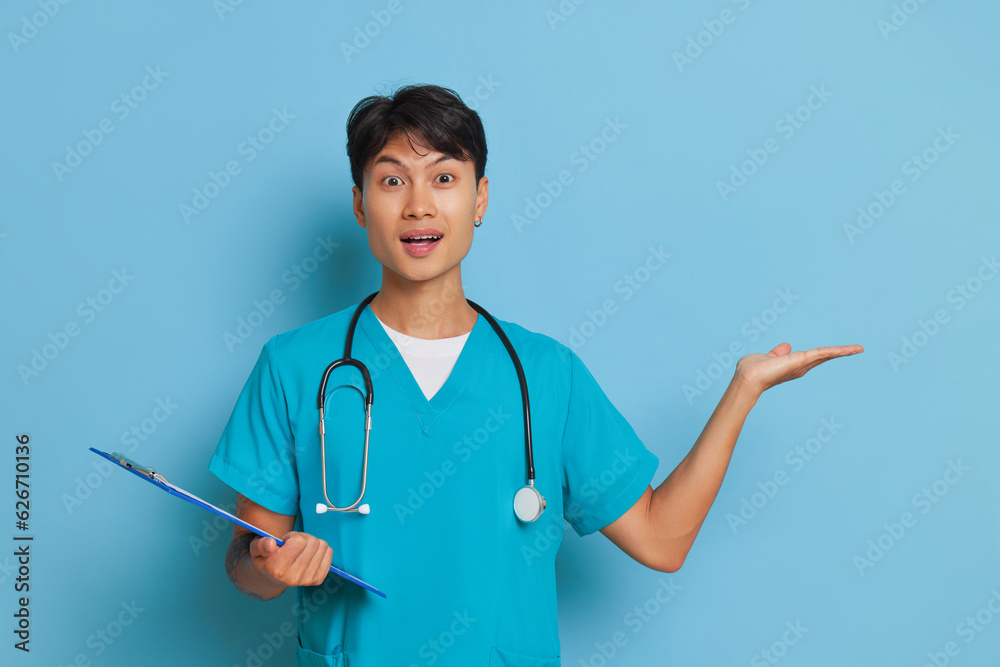 Young cheerful male doctor in turquoise uniform with stethoscope on neck stands with clipboard in his hands, professional people concept, copy space Young cheerful male doctor in turquoise uniform with stethoscope on neck stands with clipboard in his hands, professional people concept, copy space