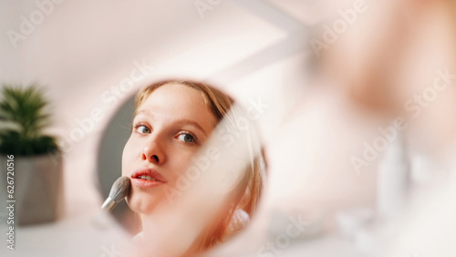Fototapeta Naklejka Na Ścianę i Meble -  Facial makeup. Cosmetic tool. Visage beauty. Woman applying natural foundation on clean face skin using powder brush in mirror reflection on light copy space.