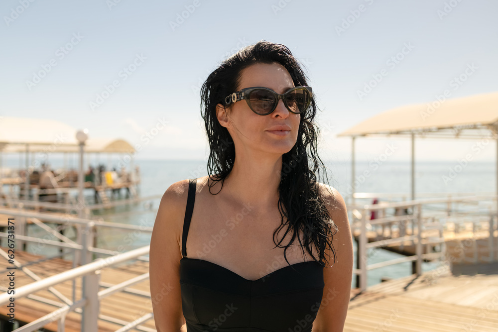 Beautiful long hair fashion brunette woman posing on the wooden pier on the beach. Dressed in a black swimsuit with straps and sunglasses. Beautiful long hair fashion brunette woman posing on the wooden pier on the beach. Dressed in a black swimsuit with straps and sunglasses.