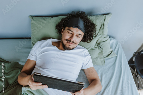 latin young man waking up and holding a tablet in bed at home in Mexico Latin America, hispanic people 