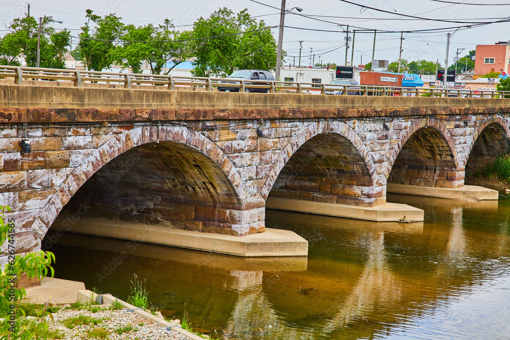 Fototapeta premium Cars crossing over old stone bridge with four arches on underside and flowing river